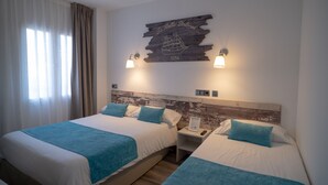 Ein Hotelzimmer mit zwei Betten, jeweils mit blauer Decke, weißer Bettwäsche und passenden blauen Kissen. Ein hölzerner Kopfteil zeigt eine Illustration eines Schiffes und den Schriftzug "La Bella Del Mar 1954". Auf jeder Seite des Kopfteils befinden sich zwei Wandleuchten.
