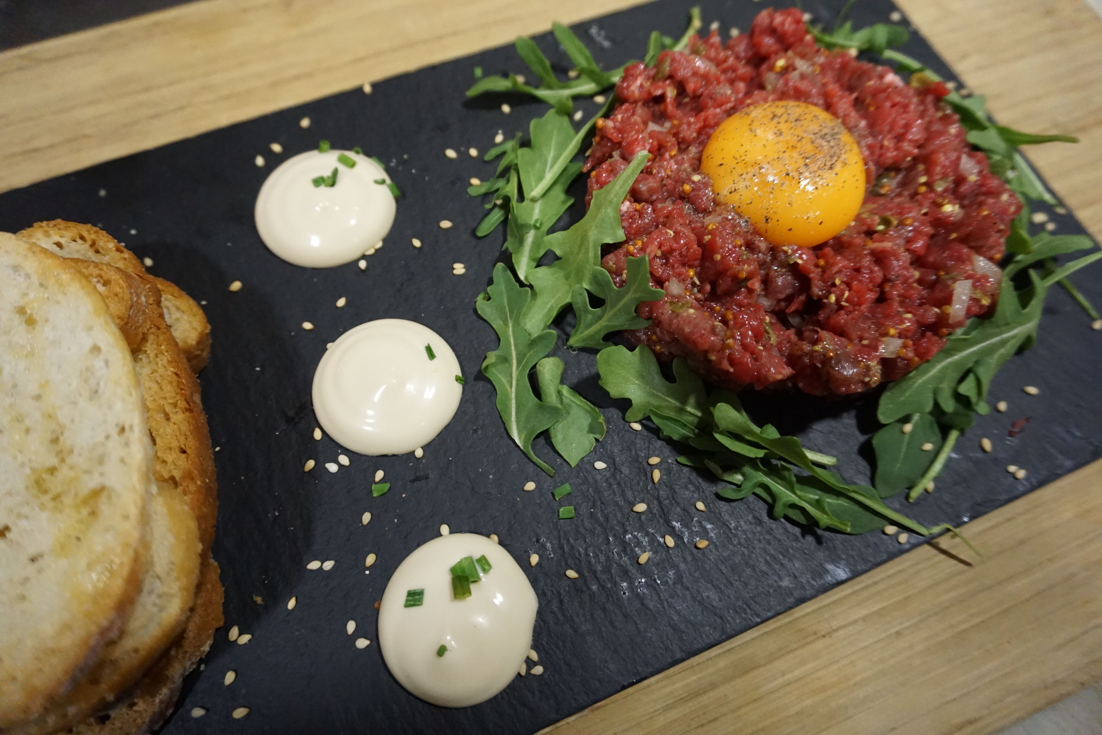 A dish of steak tartare with a raw egg yolk, arugula, and condiments on a slate plate.