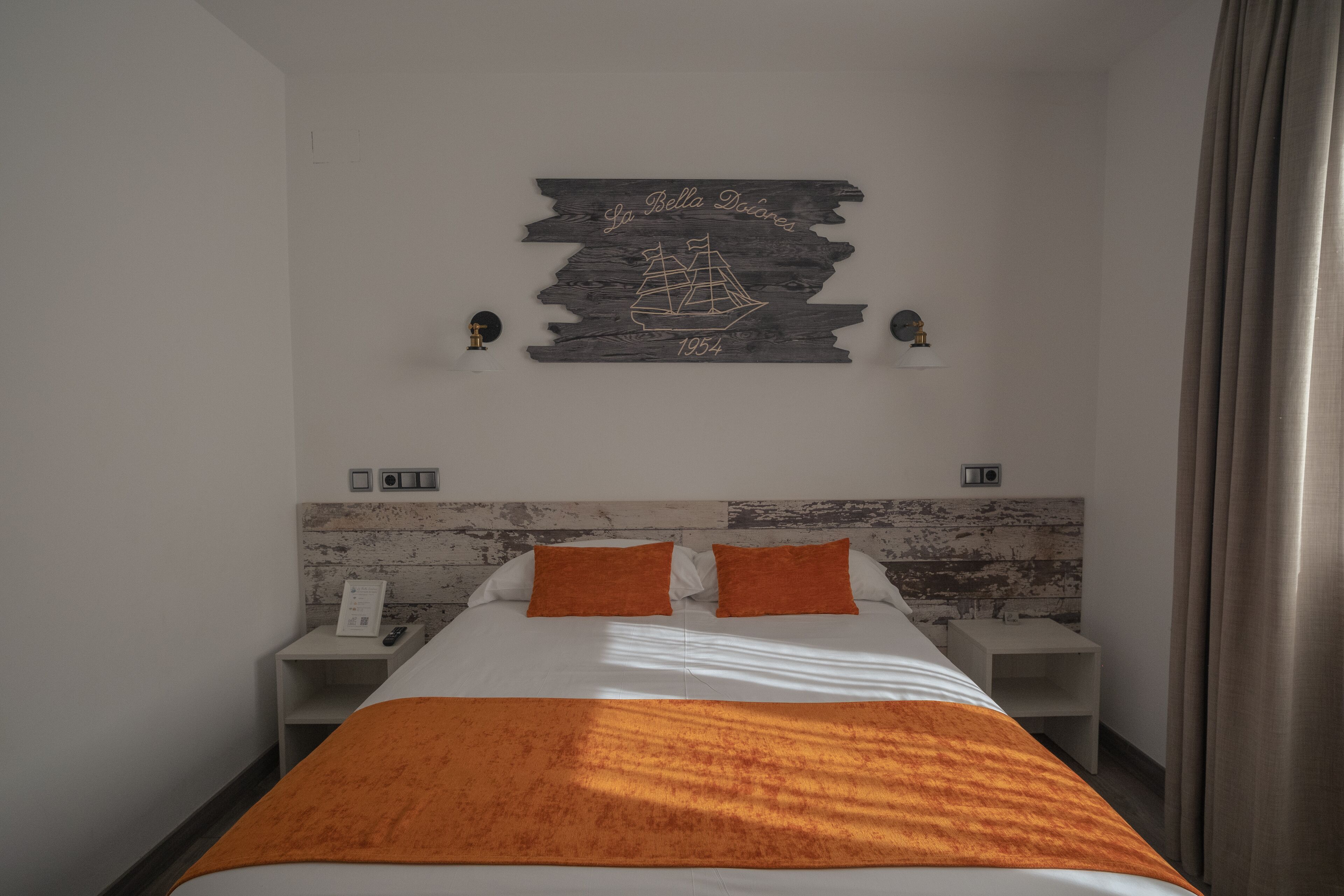 Une chambre d’hôtel comprenant un lit, deux tables de chevet, une tête de lit en bois, une œuvre d’art accrochée au mur et un rideau.