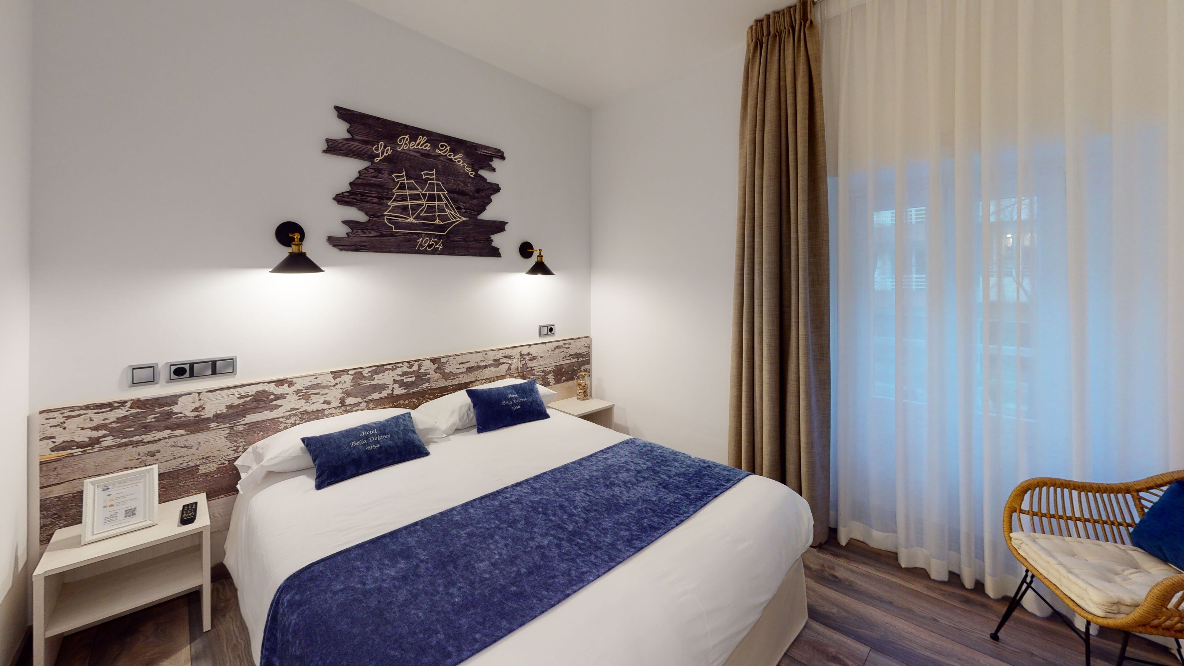 A hotel room with a large bed, a headboard with a distressed wood design, a wall-mounted ship decoration, two bedside lamps, a nightstand with a remote control, and a wicker chair.