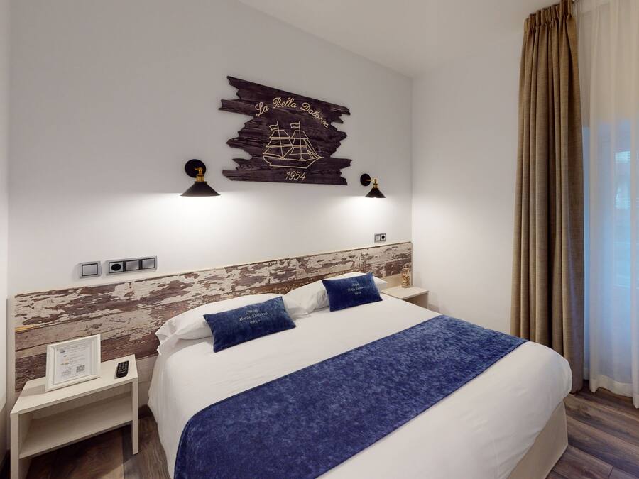 A hotel room with a large bed, a headboard with a distressed wood design, a wall-mounted ship decoration, two bedside lamps, a nightstand with a remote control, and a wicker chair.
