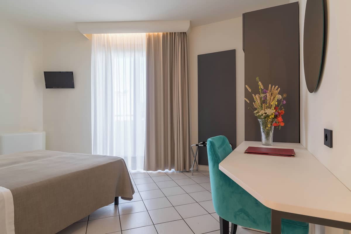 Une chambre d’hôtel comprenant un lit, un bureau, une chaise, une télévision, une fenêtre avec des rideaux et un vase avec des fleurs.