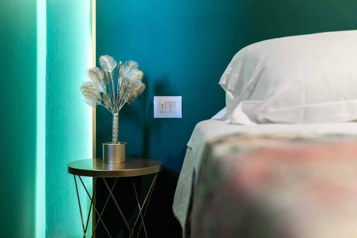 A room with teal walls, a bed with white bedding, a small round table with a metallic vase, and a wall switch.