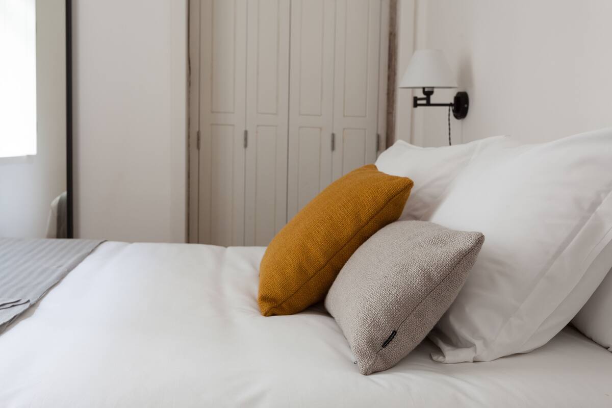A neatly made bed with white and mustard pillows, a white wall, and a closed door in the background.
