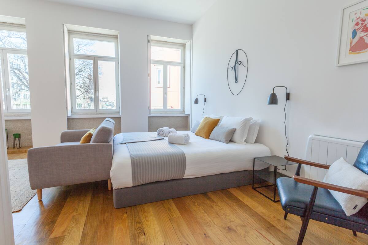 A modern bedroom with a large bed, a grey sofa, a wooden floor, and a window.