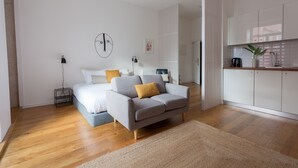A modern bedroom with a grey sofa, a bed with white bedding, a small bedside table with a lamp, and a kitchen area with white cabinets and a plant. - City View - Santa Catarina (Porto)