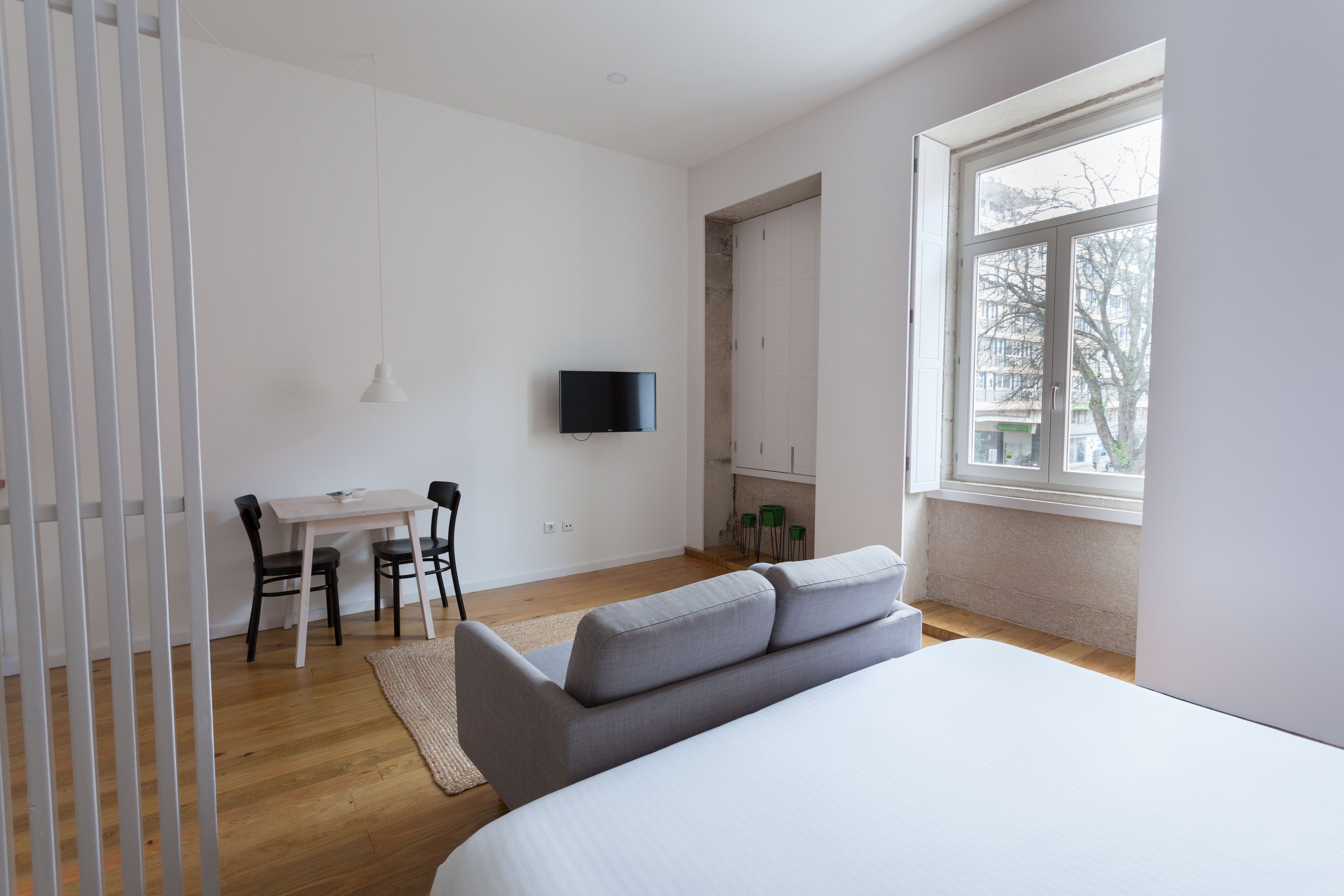 A modern bedroom with a bed, a TV mounted on the wall, a dining area with a table and chairs, and a window providing natural light.