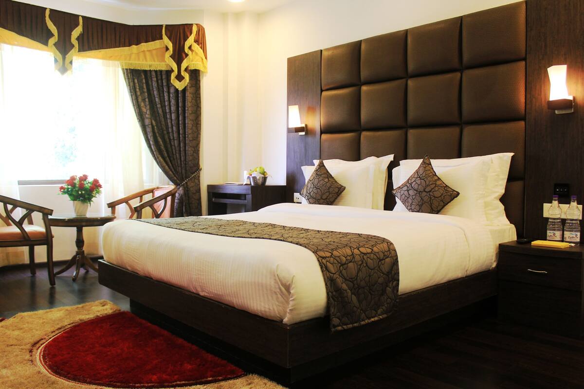 Quarto de hotel com uma cama grande, uma mesa de cabeceira, uma cadeira, uma pequena mesa com um vaso de flores e uma janela com cortinas.