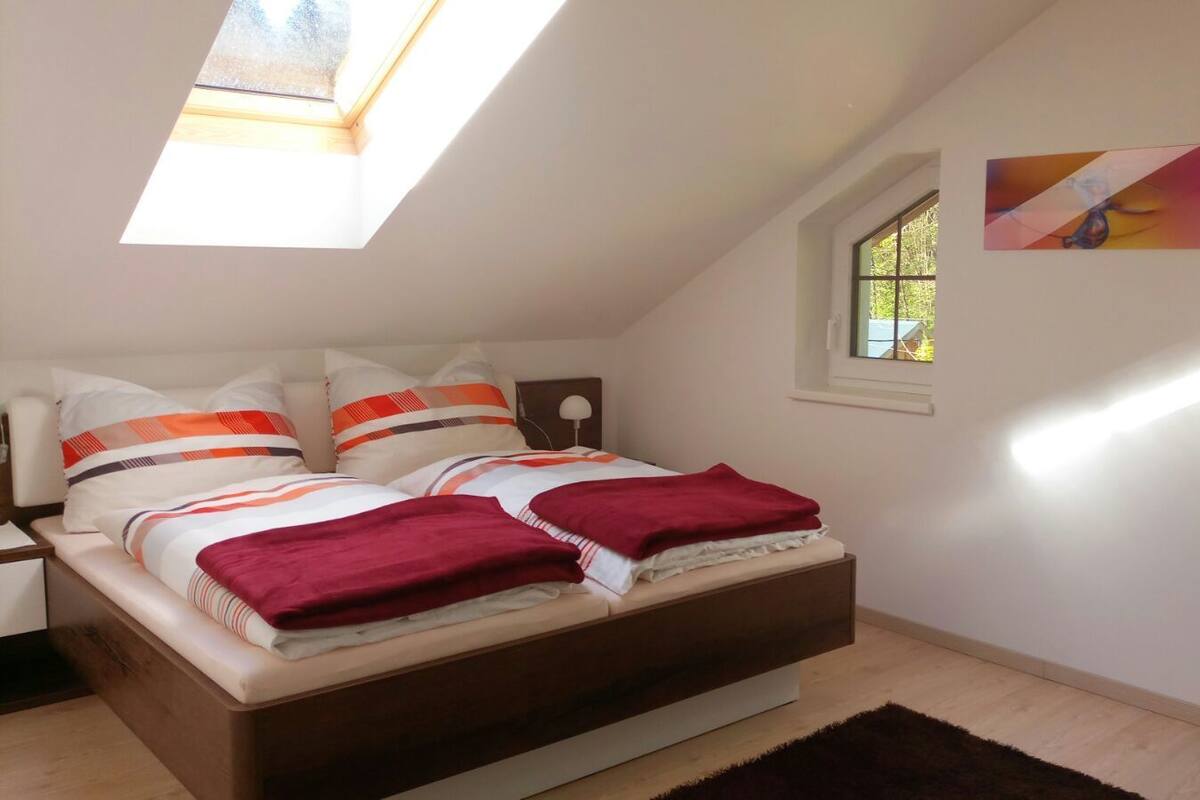 Une chambre avec une lucarne, un lit agrémenté de plusieurs oreillers, une table de chevet, une fenêtre donnant sur un beau paysage et un tableau accroché au mur.