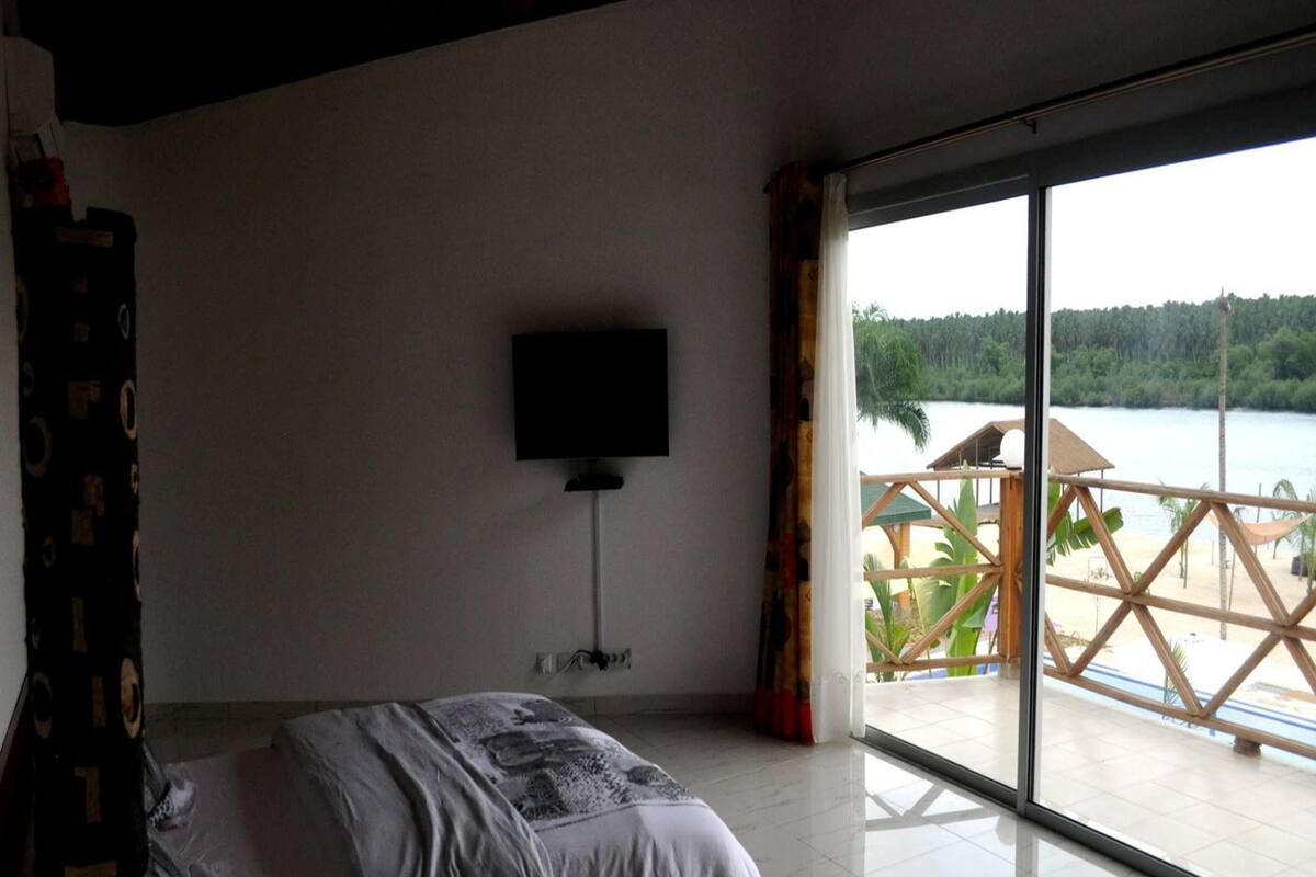 A bedroom with a bed, a TV on the wall, and a large window overlooking a deck with a view of a lake and trees.
