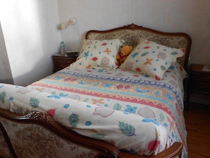 A bed with a floral-patterned bedspread and two pillows, a bedside table with a bottle, and a nightstand with a lamp. - Chez Colombine (Chaponost)