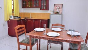 Un comedor con una mesa de madera dispuesta para cuatro personas, rodeada de sillas. Se ve una cocina con armarios rojos y un refrigerador pequeño al fondo.