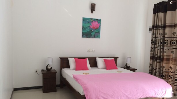 A neatly arranged bedroom with a wooden bed, white and pink bedding, a small bedside table with a lamp, and a wall-mounted light fixture. - Lo hostel (Weligama)