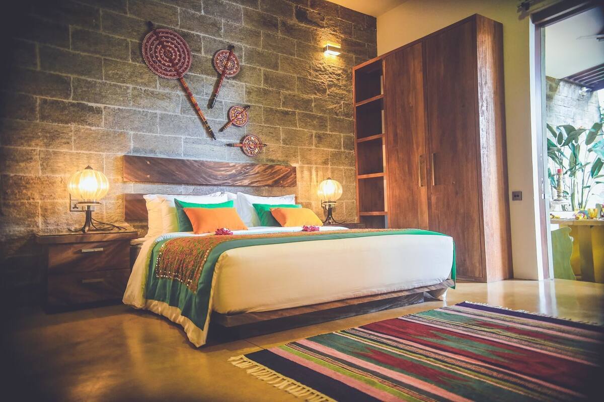 Une chambre avec un mur en pierre, une tête de lit en bois, deux lampes de chevet, un lit avec des coussins colorés, un tapis à rayures et une armoire en bois.