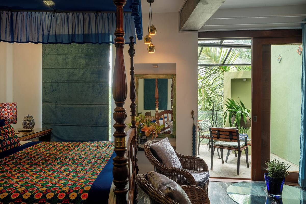 Une chambre avec un lit à baldaquin, un coin repas avec des meubles en osier et une vue sur un espace extérieur verdoyant.