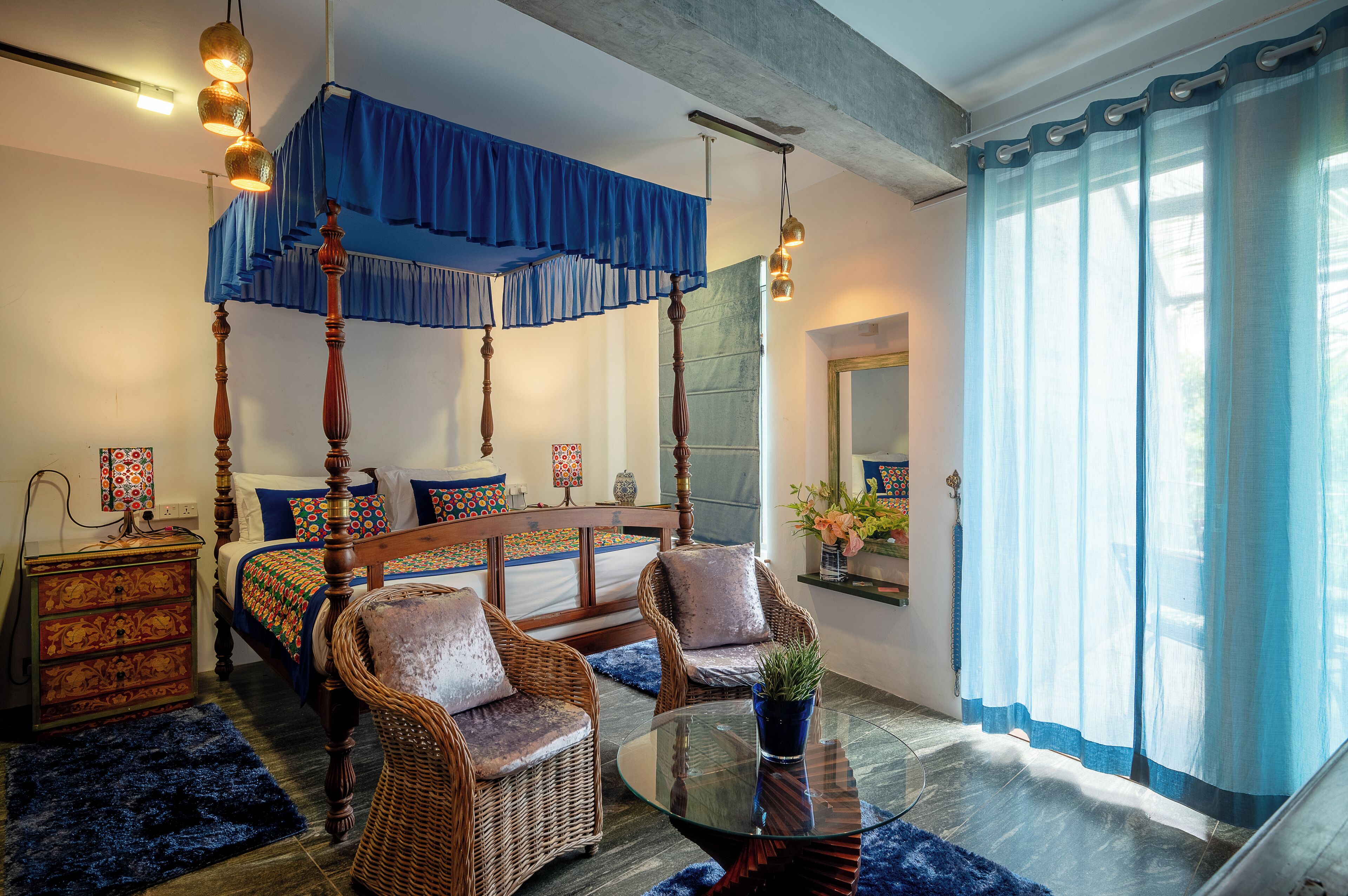 A four-poster bed with a blue canopy, a glass coffee table, wicker chairs, and a blue rug in a room with a large window and curtains.