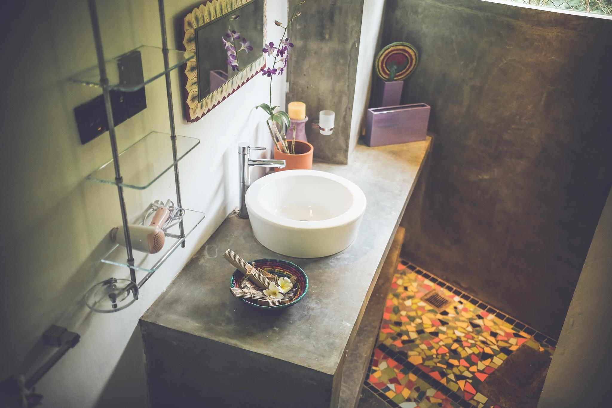 A bathroom with a large white sink, a glass shelf with toiletries, a mirror with a decorative frame, and a colorful mosaic floor.