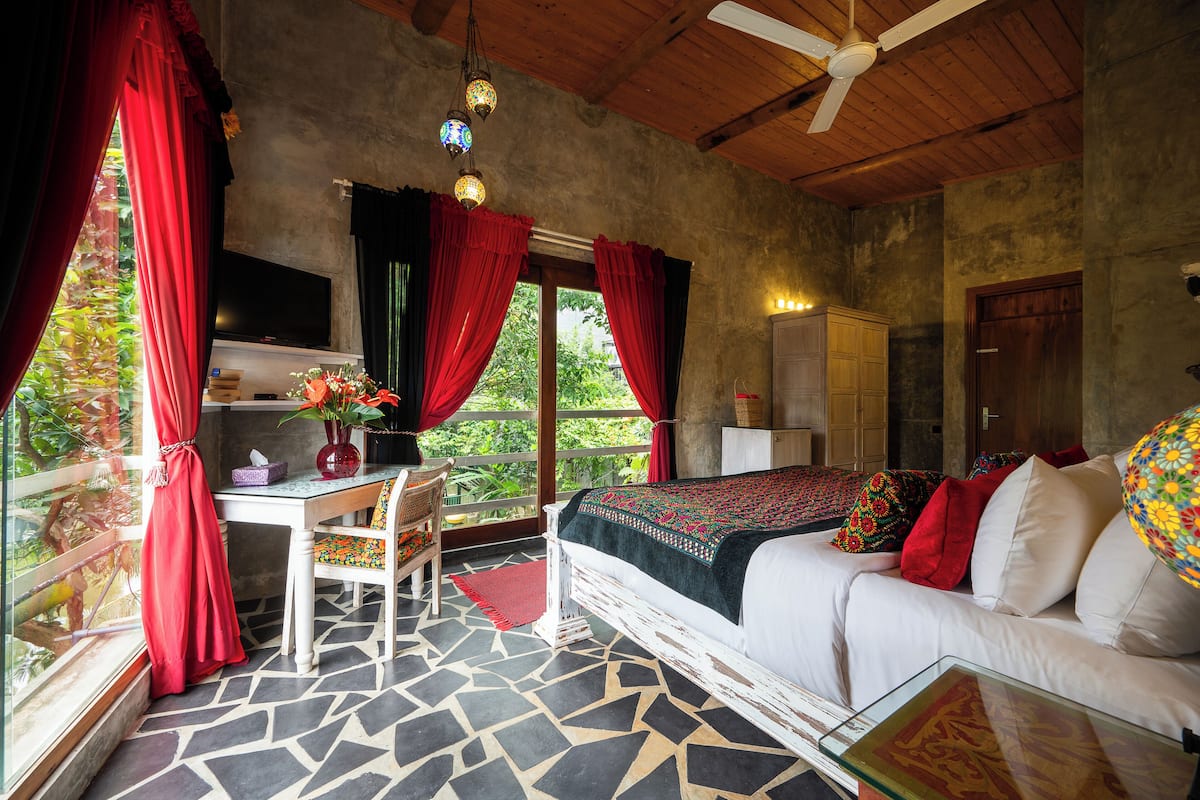 Une chambre avec un lit, un bureau et une chaise. Il y a une fenêtre avec des rideaux rouges, un ventilateur de plafond et un téléviseur fixé au mur.