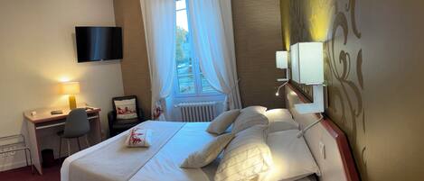 A hotel room with a bed, a desk with a chair, a TV mounted on the wall, and a window with curtains.