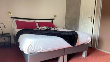 Una cama bien hecha con ropa blanca y roja, una manta de color negro y una almohada estampada. Hay una mesita de noche pequeña con una lámpara y un enchufe. La habitación tiene un diseño sencillo y limpio, con una puerta a la derecha y una pared lisa.