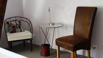 Una habitación pequeña con una cama individual, una silla de madera con cojín, una mesita redonda con lámpara y un cubo de basura rojo.