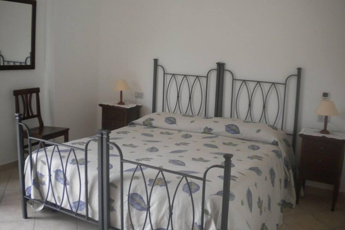 Une chambre simple et épurée, avec un lit en métal, une seule chaise et deux tables de chevet équipées de lampes.