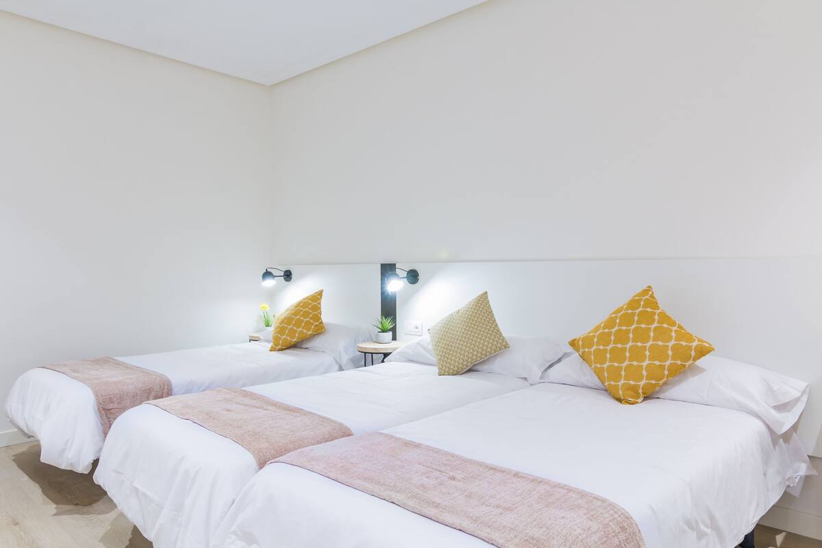 A hotel room with two beds, each with white bedding and a pink throw blanket. There are yellow patterned pillows and a small table with a plant.