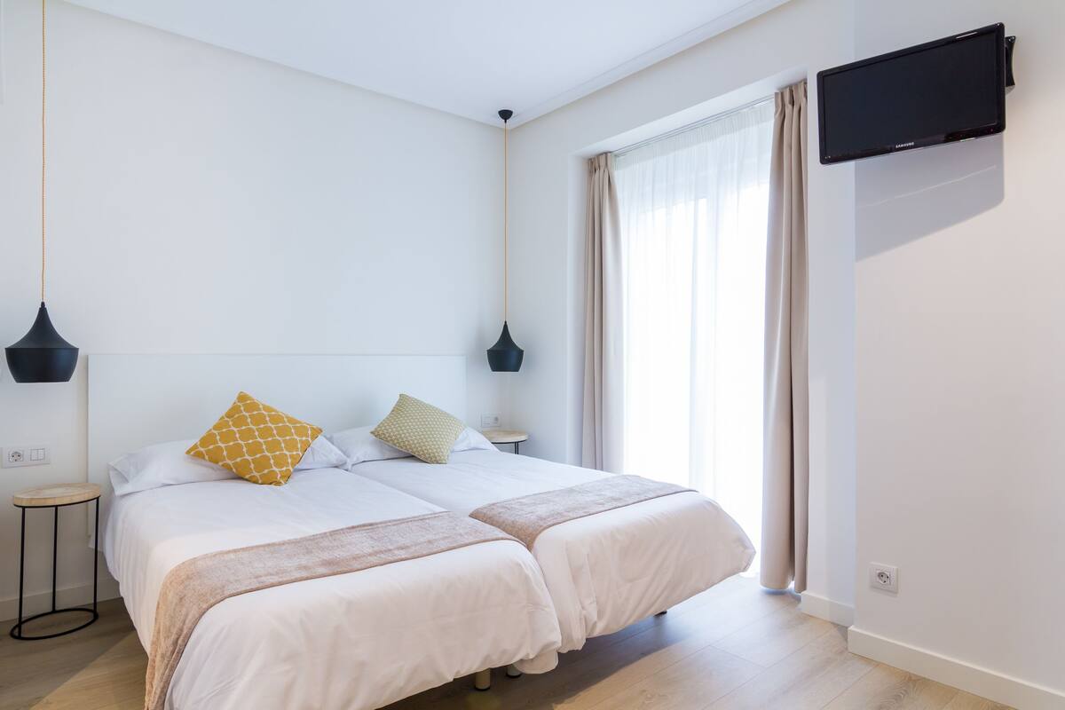 A modern hotel room with two beds, a flat-screen TV, and a large window with curtains.