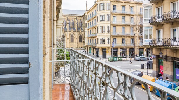 A balcony with wrought iron railings overlooking a street with buildings, a pedestrian crossing, and a parked car. - Pensión BUENPAS (San Sebastián)