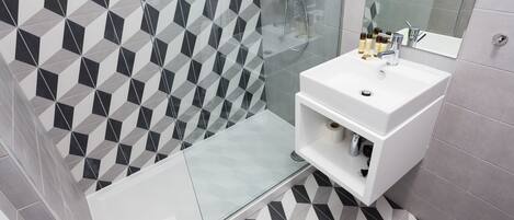 Un baño moderno con un diseño geométrico de azulejos, un box de ducha de cristal, un lavabo blanco con artículos de aseo y un estante empotrado.
