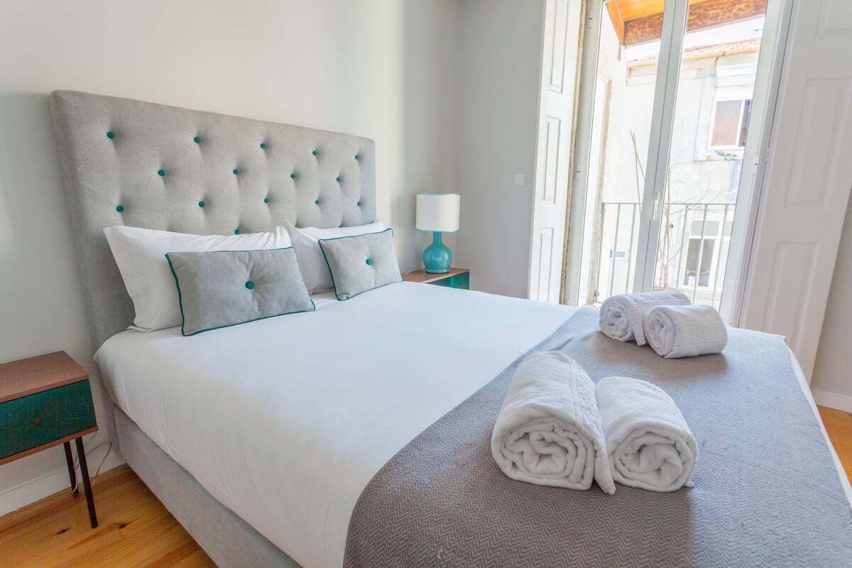 A neatly made bed with a grey headboard and pillows, a wooden nightstand, and a window with a view of buildings.