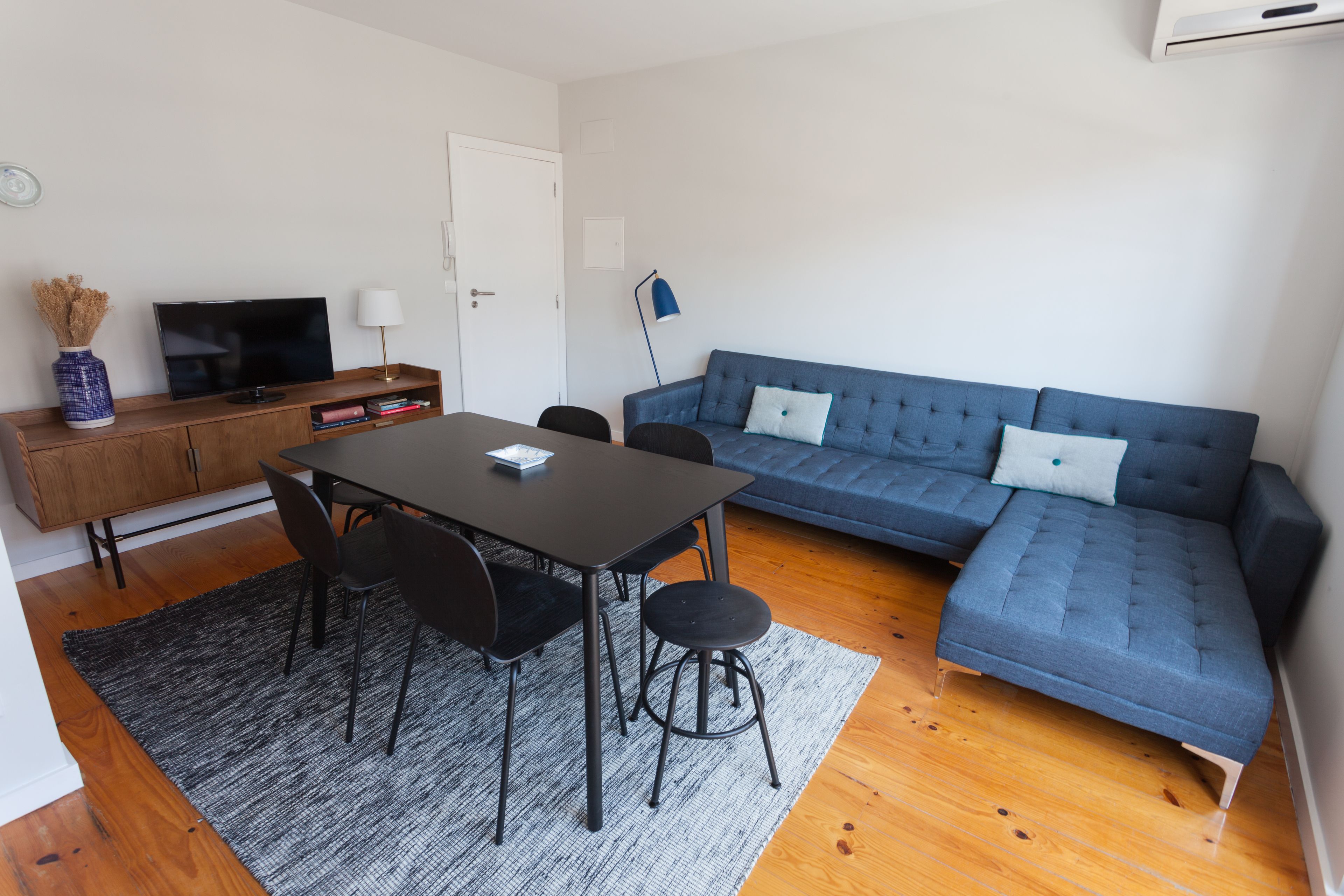 A modern living room with a sectional sofa, a dining table with chairs, a TV on a wooden cabinet, and a lamp.
