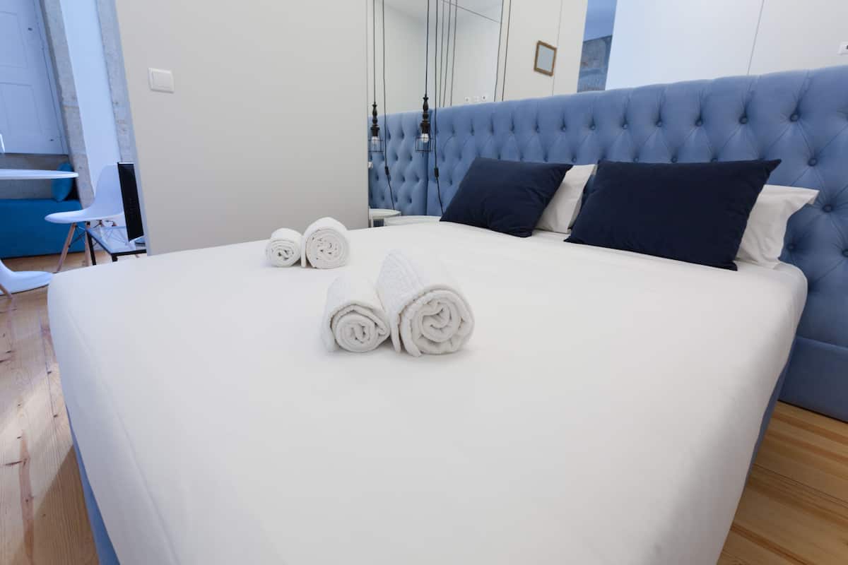 A neatly made bed with white linens and two rolled towels, a tufted headboard, and a small bedside table.