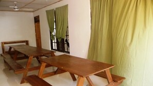 A room with three wooden tables and benches, a ceiling fan, and a window with green curtains. - Palugaha Pilgrim Resort (Kataragama)