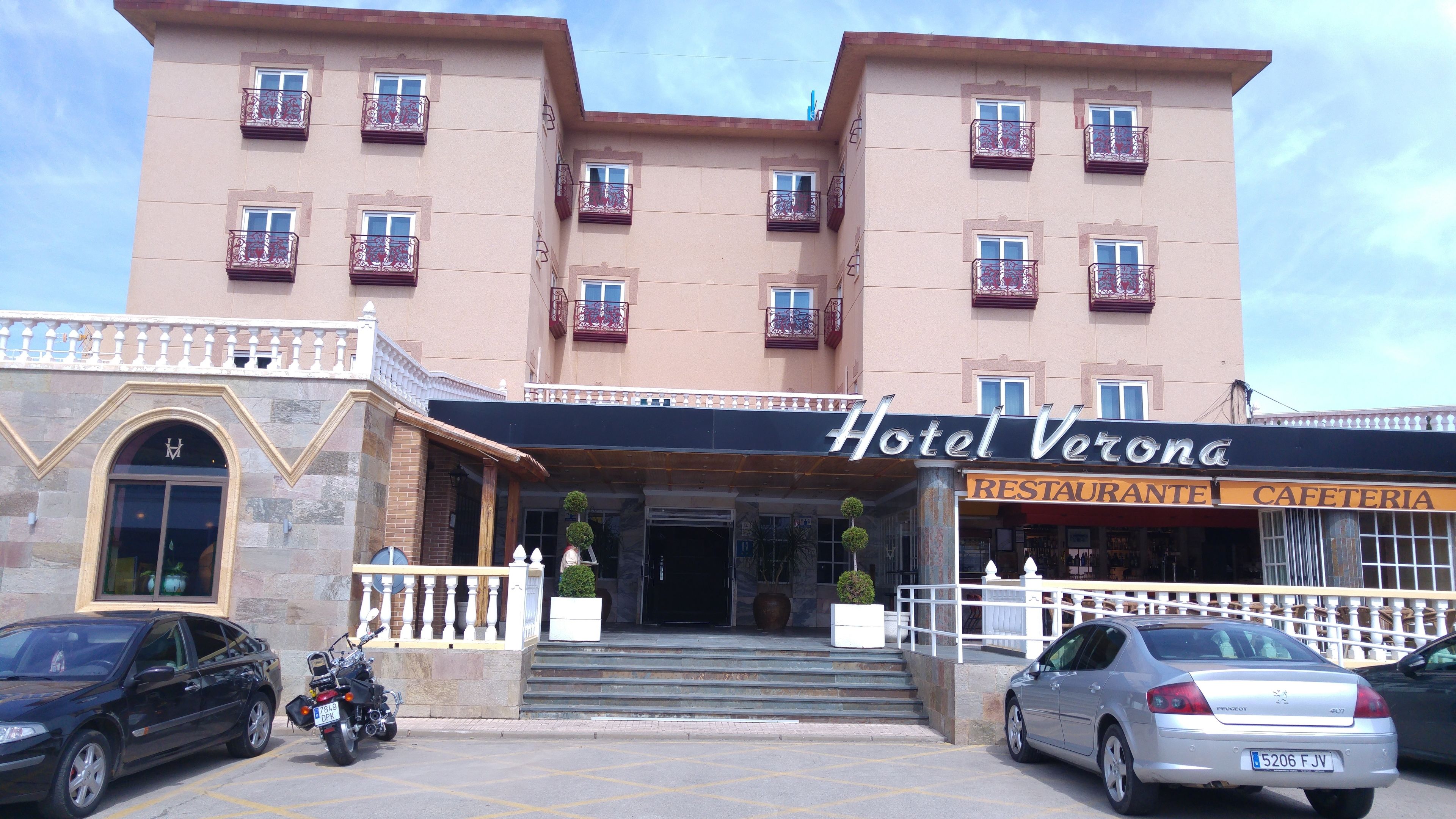 Un hotel con una entrada destacada, un restaurante y un estacionamiento con coches y una motocicleta.