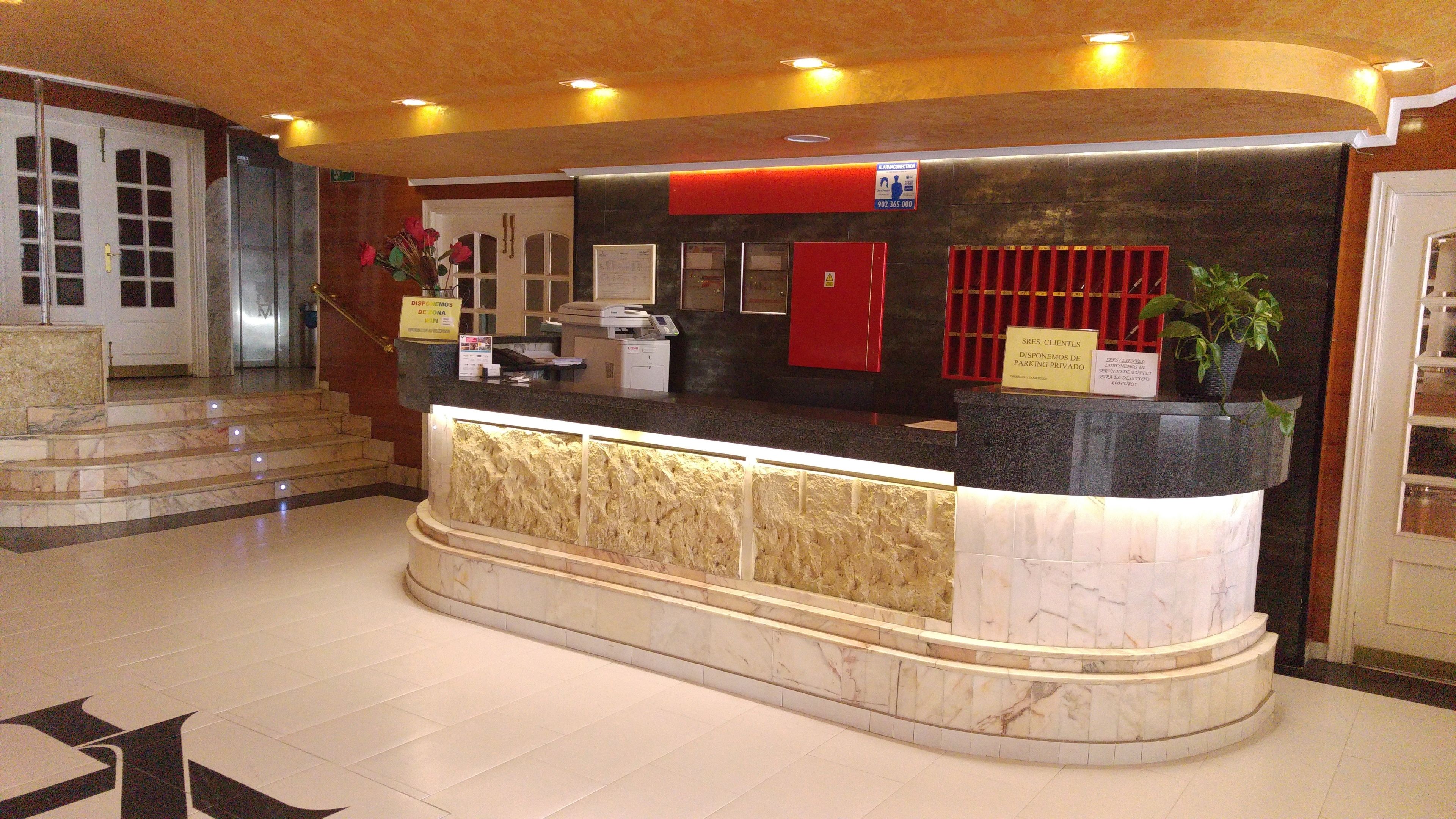Un comptoir d’accueil d’hôtel avec un plan de travail en marbre, un panneau d’information rouge et un escalier menant à un étage supérieur.