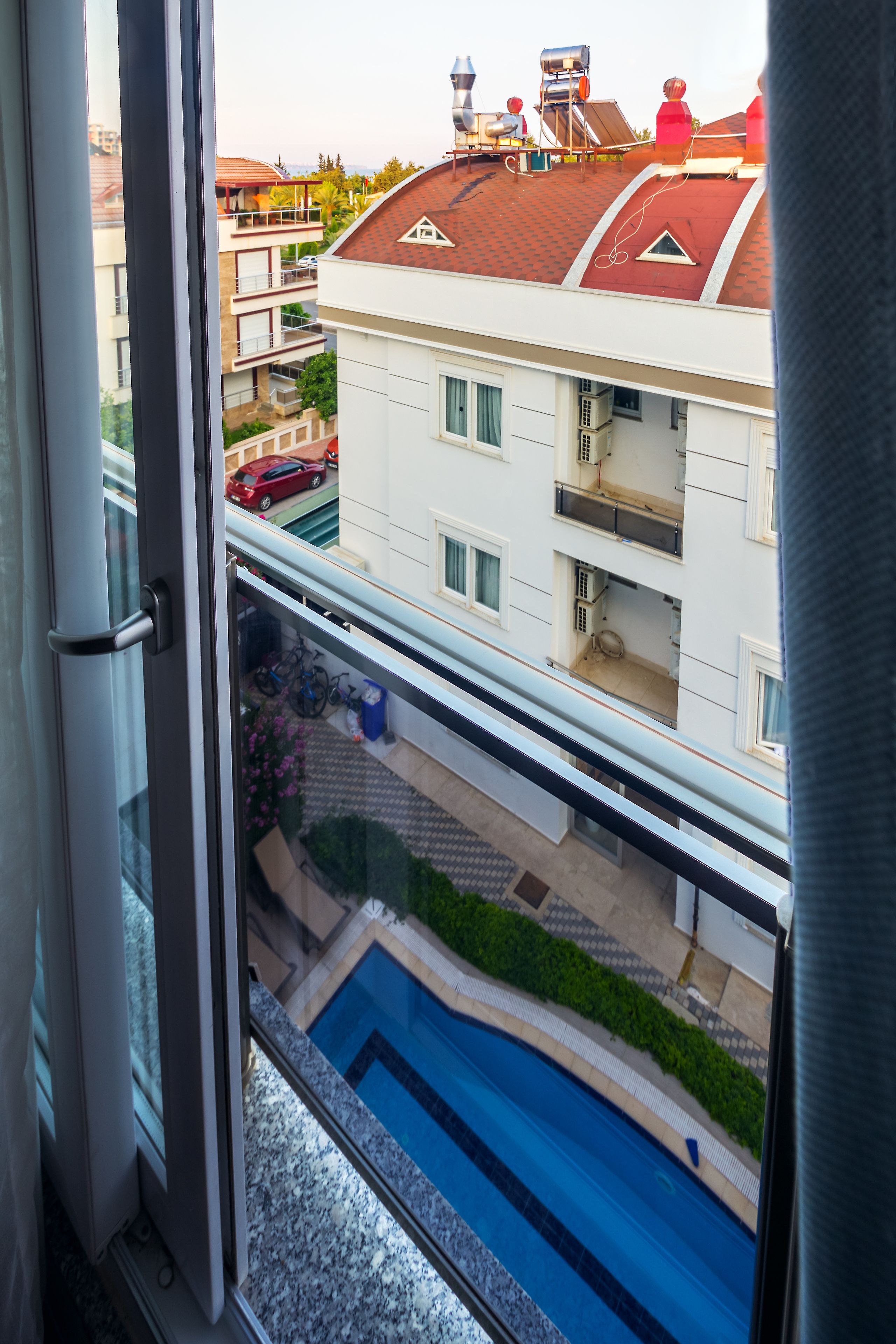 Une vue depuis une fenêtre donnant sur une piscine, une voiture rouge et un immeuble à plusieurs étages doté de balcons.