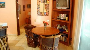 A dining area with a round wooden table, two chairs, a vase with flowers, a television on a wooden shelf, and a bulletin board with various pictures. - Casita Bendon by Las Palmas (Bucerías)