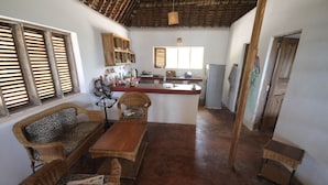 Un vaste espace de vie comprenant une table à manger en bois, deux canapés en osier et un coin cuisine équipé d’un réfrigérateur et de meubles de rangement en bois.