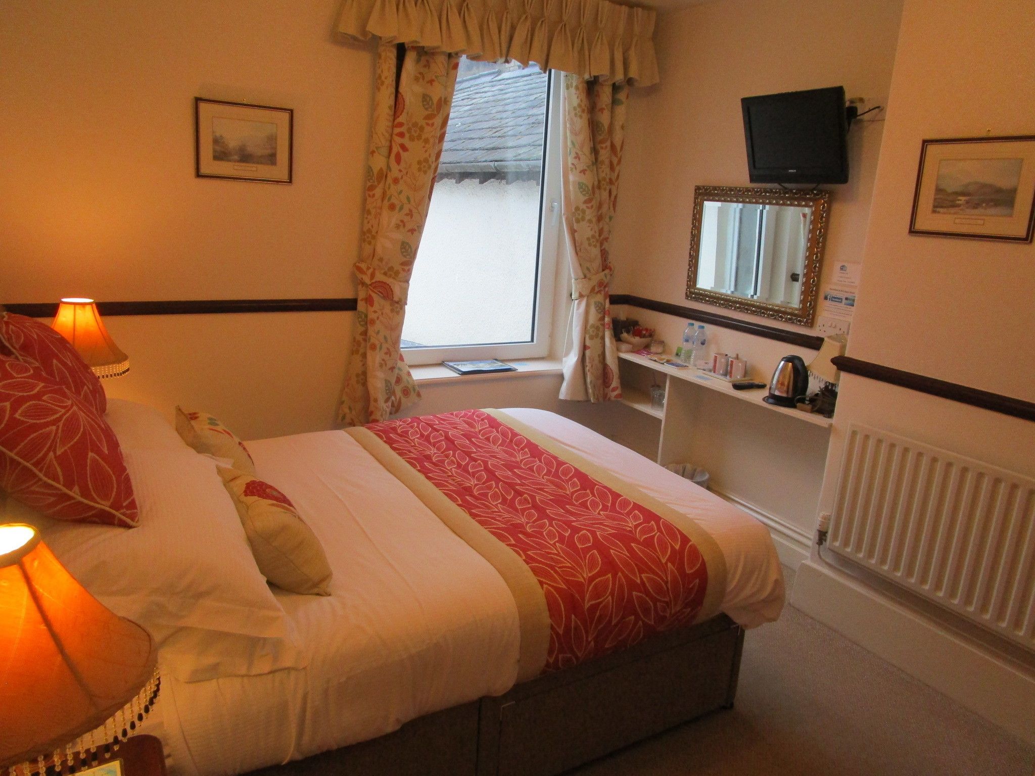 A hotel room with a bed, bedside lamps, a window with curtains, a mirror, a television, and a radiator.