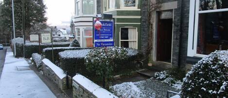 Una calle residencial con setos cubiertos de nieve y un letrero que indica 'No hay habitaciones disponibles' para una propiedad llamada 'Brookfield Walkers Welcome'.