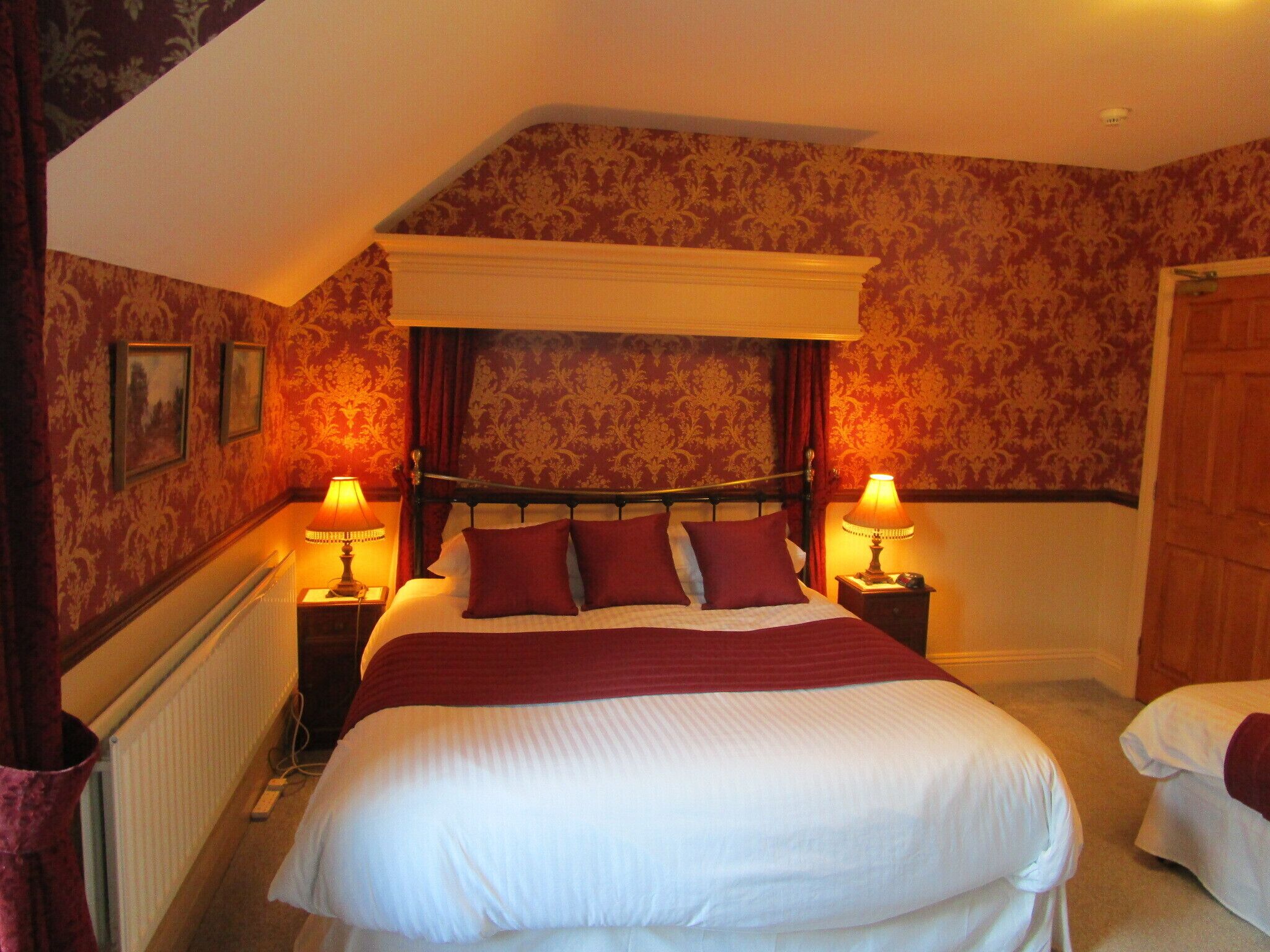 A bedroom with a large bed, two bedside tables with lamps, patterned wallpaper, and a wooden door.