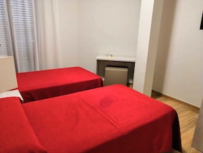 Una habitación con dos camas cubiertas con sábanas rojas, un escritorio pequeño con una silla y una ventana con persianas.