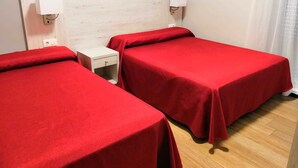 Una habitación con dos camas individuales, cada una con una colcha roja, una mesita de noche pequeña con una lámpara y un cabecero blanco.