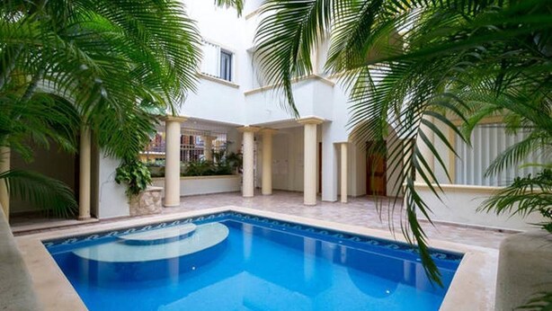 A swimming pool with a tiled deck, surrounded by palm trees and a white building with columns. - Arcoiris Dreams by Playa Paradise (Playa del Carmen)