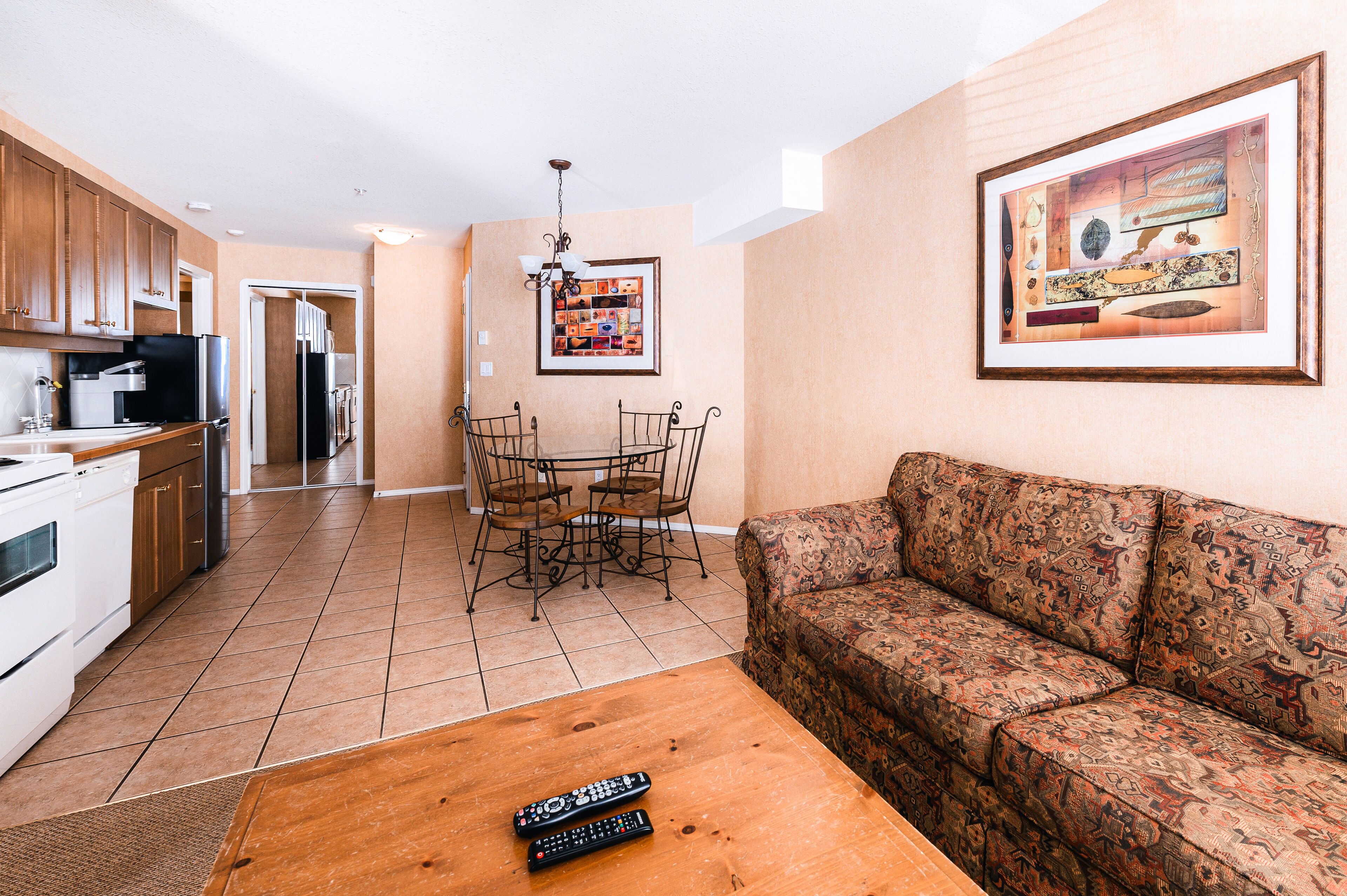A living room with a sofa, a coffee table with remotes, a dining area with chairs, and a kitchen with cabinets and appliances.