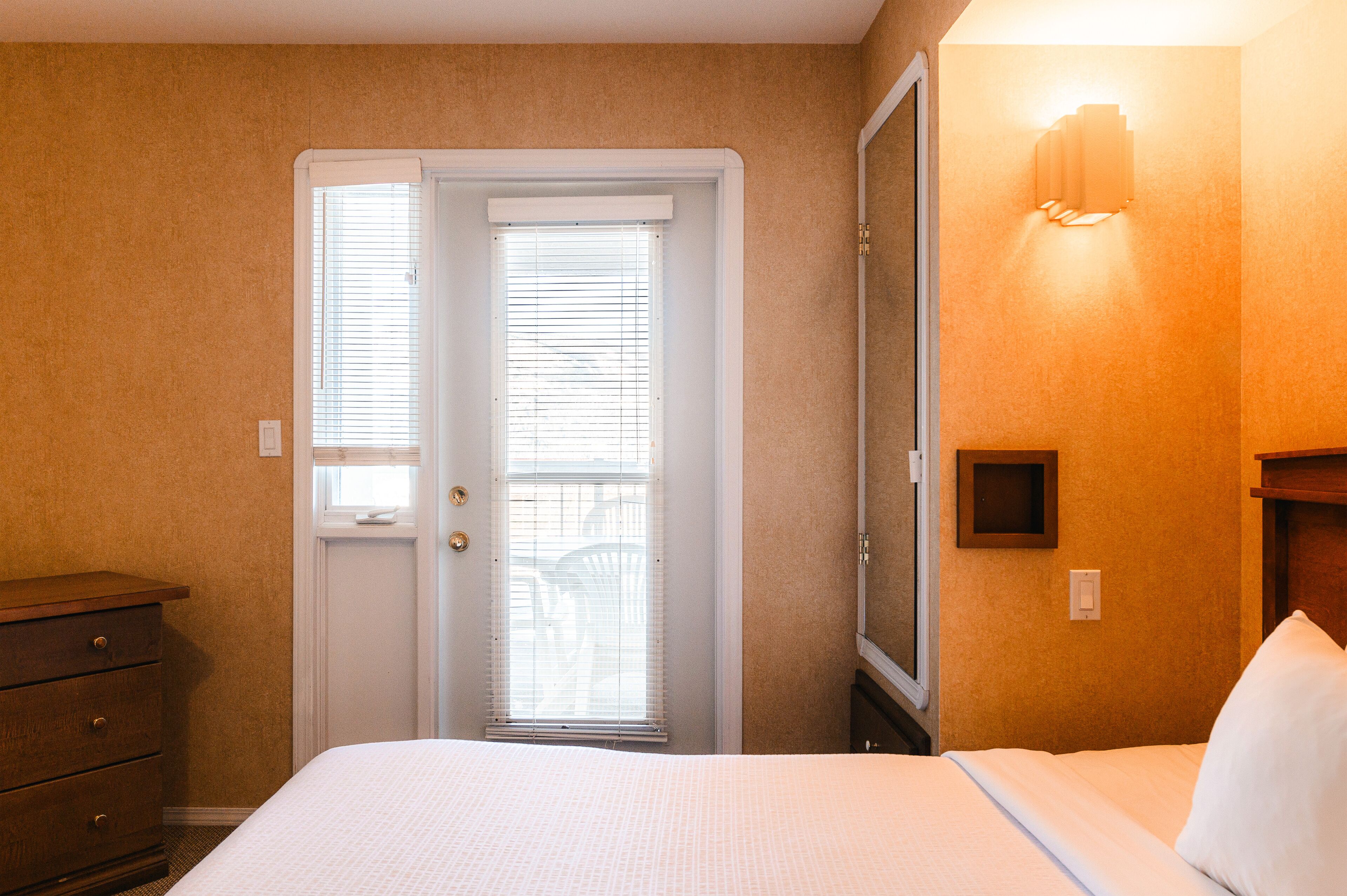 Una habitación de hotel con cama, cómoda, puerta con persianas y una lámpara fijada en la pared.