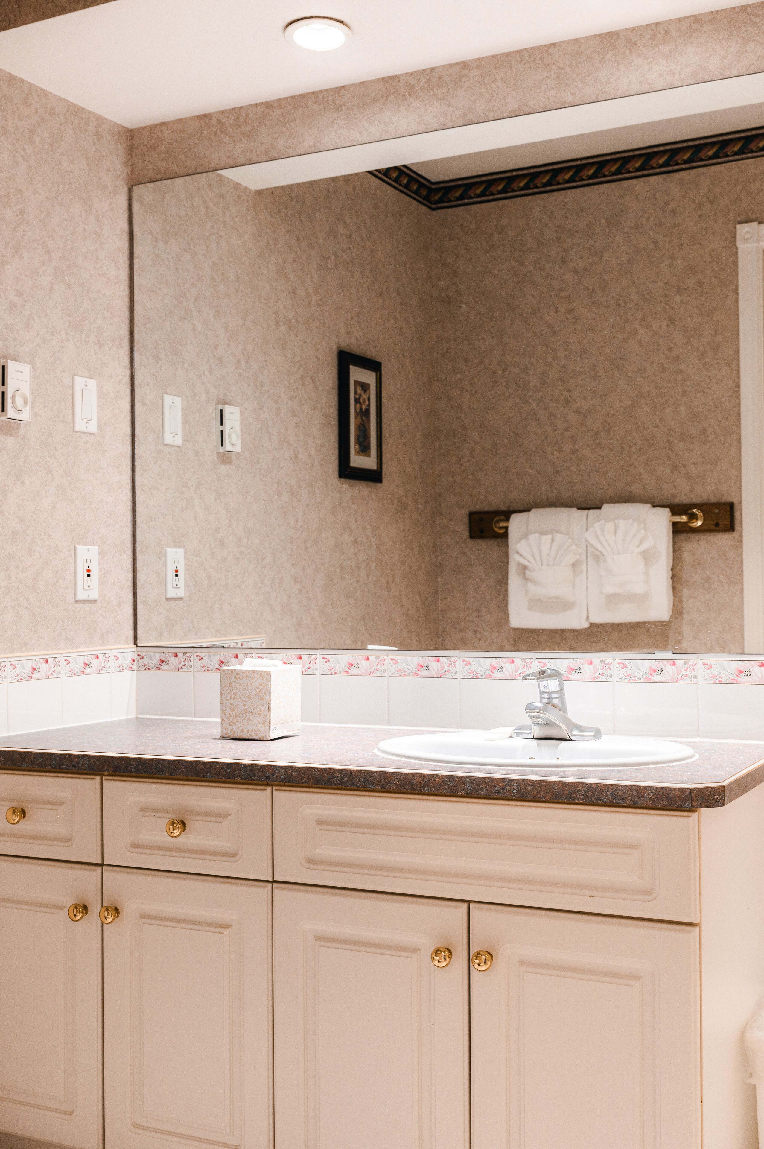 A bathroom with a beige tiled wall, a white sink, and a mirror above it. There are two towel racks with white towels, and a small framed picture on the wall.