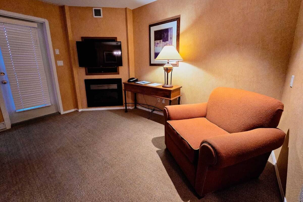 Una habitación de hotel con un sillón naranja, un escritorio con lámpara, televisión y chimenea.
