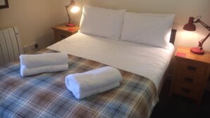 Cama bem arrumada com colcha xadrez, duas luminárias de cabeceira e uma pequena cômoda de madeira com gavetas.